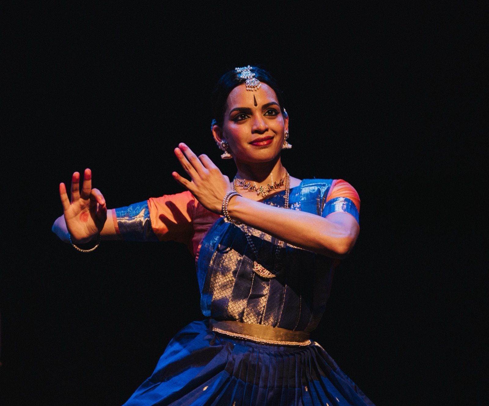 Vertika on stage forming a hasta mudra near her face, in a blue and orange costume, against a dark background.