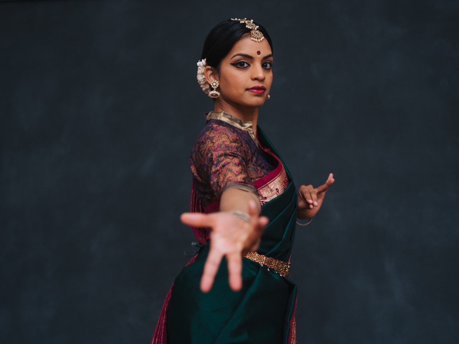 Vertika in a green and maroon silk saree, one hand extended toward the camera, against a dark grey backdrop.