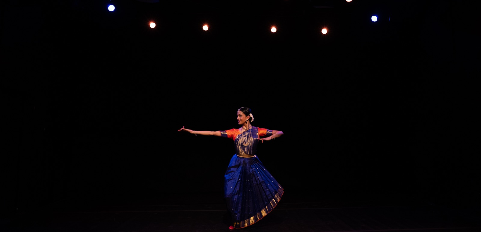 Vertika performing bharatanatyam alone on a dark stage under spotlights, in a blue and orange costume.
