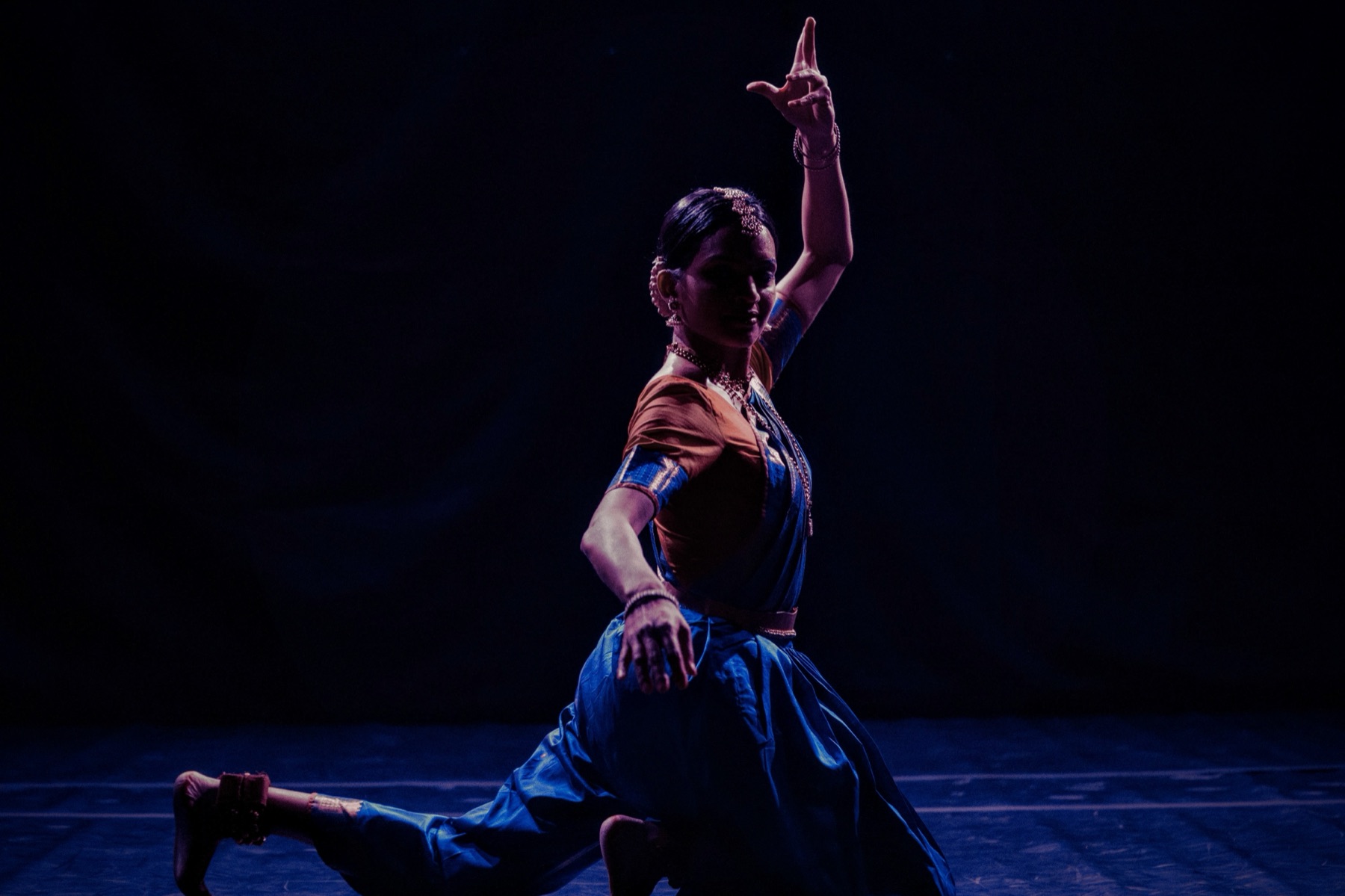 Vertika mid-lunge on stage, arm raised, in a blue and orange costume.